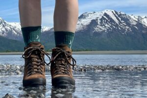 A hiker stands shallow water in leather books and Darn Tough socks. There is a river behiind her, with snow capped mountains in distance.