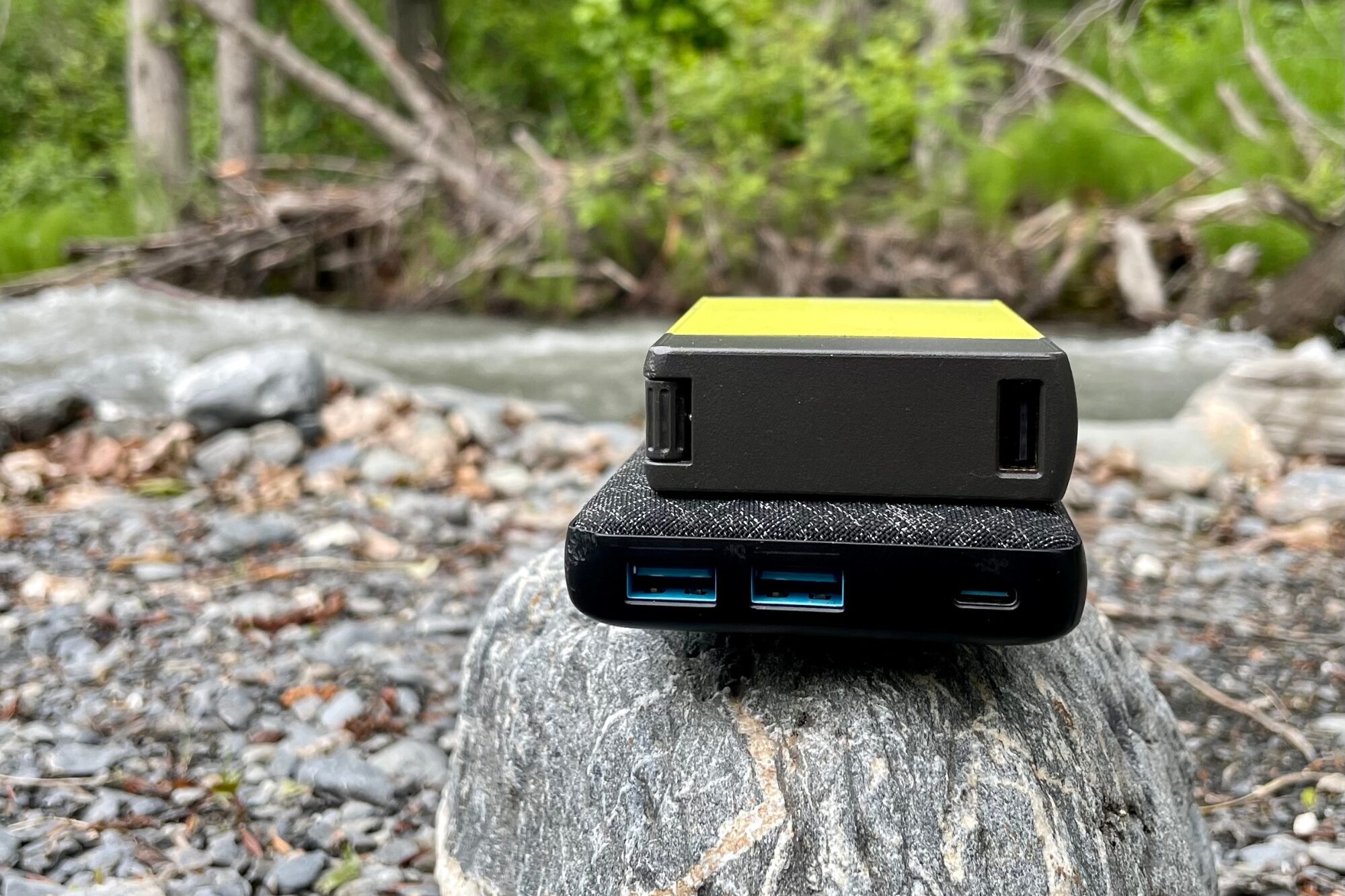Two power banks are stacked on top of each other, showing the different ports and sizes. They're sitting on a stone in front of a river.