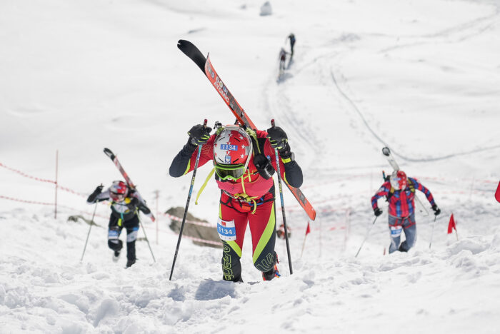A skimo racer looking down, with skis on backpack. 