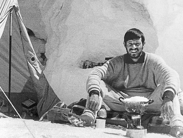 Guenther Messner in a black and while photo, sitting by a tent, with icce behind him and a cooking stove in front. 