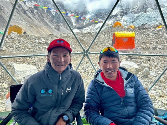 Lakpa and Mingma sits in EBC mess tent, mountains behind the transparent plastic wall. 