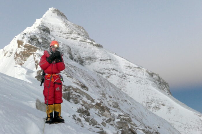 Ballingers stands on a snow and ice slope, with a gedlamp on and Everest somital pyramid in background. 