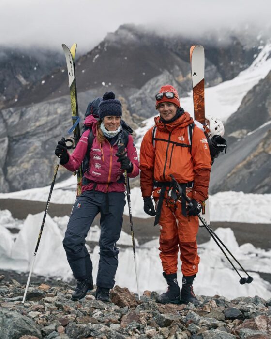 Tybor and Lafaille with skis on their backpacks at the foot of the mountain on moraine terrain.