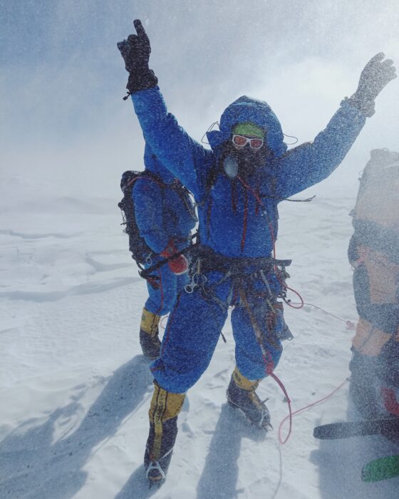 Climber on O2 raises his arms in triumph on a windy summit of Everest. 