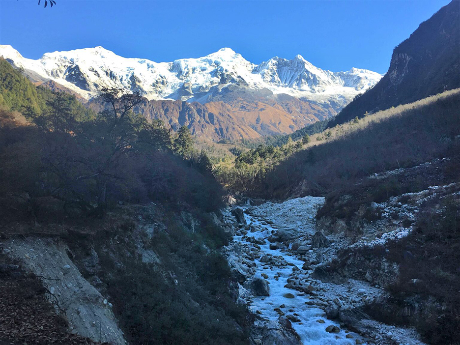 A mountain river down a valley, with the big Manaslu range in the sun in the background.