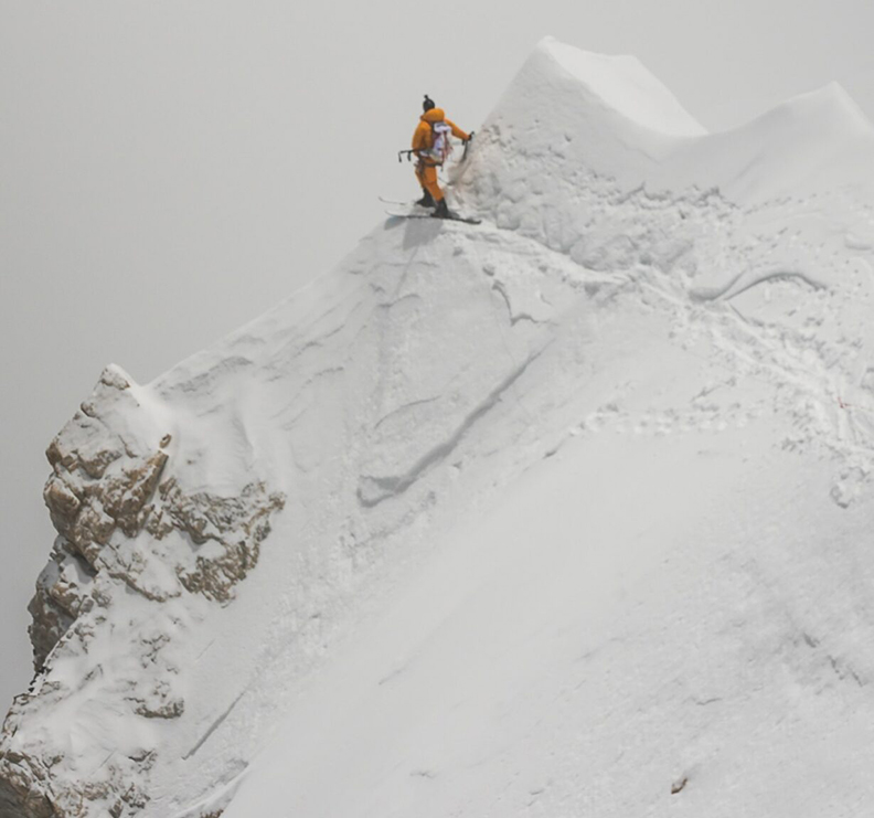 The skier peers from the edge of a nearly vertical snow cornice, in a foggy day