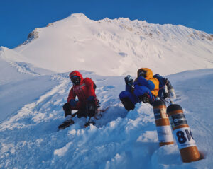 Climbers rest sitting on the snow, 2 bottles of O2 by them, and the summit ridge of Everest in background