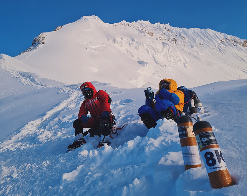 Climbers rest sitting on the snow, 2 bottles of O2 by them, and the summit ridge of Everest in background