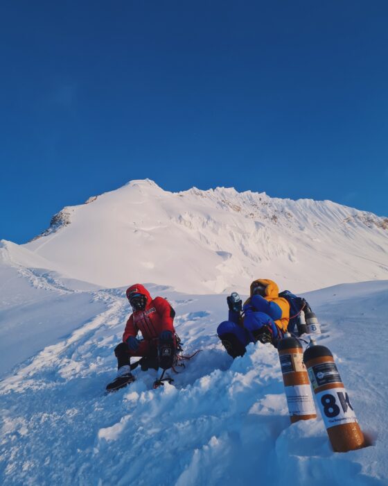 Climbers rest sitting on the snow, 2 bottles of O2 by them, and the summit ridge of Everest in background