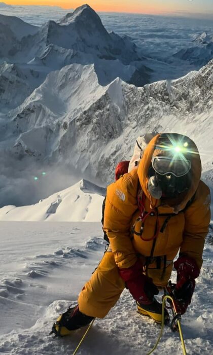 Everest climber at dawn, with headlamp on, totally covered in down suit and mask face and O2, with Makalu in background