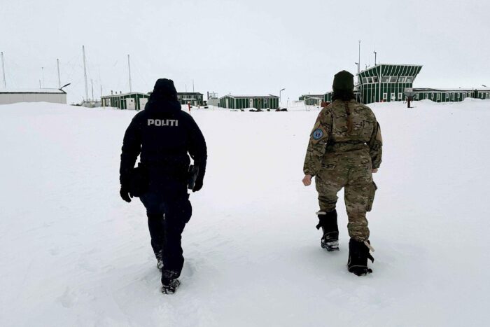 police officer walking across snow toward green buildings with camouflaged companion