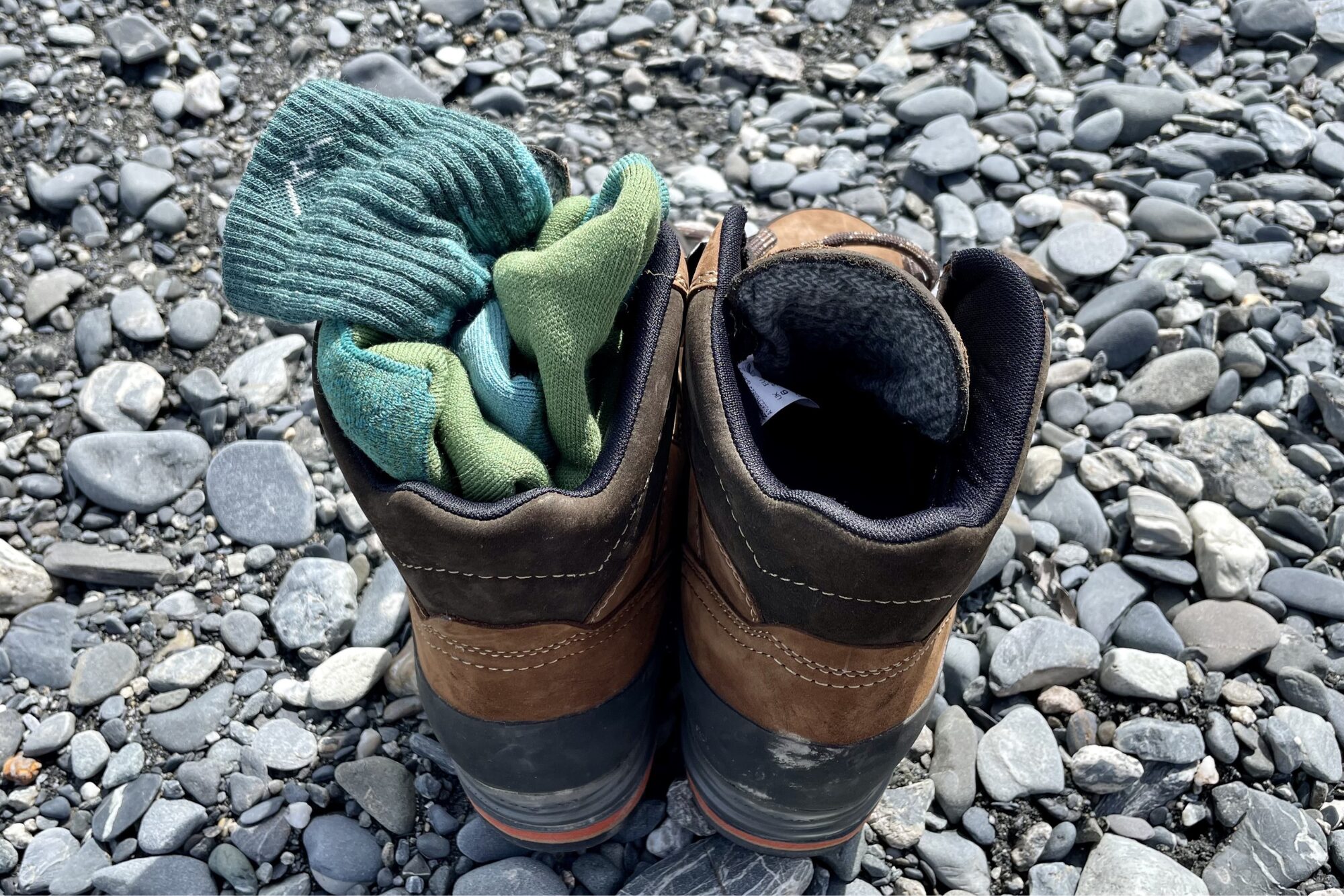 A pair of darn tough socks are stuffing into leather hiking boots sitting on a river bed. 
