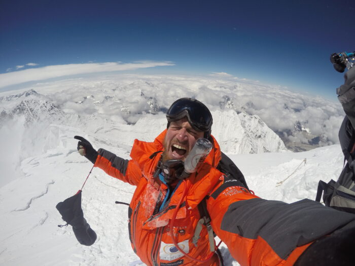 Veyt screams to the camera in triumph from the summit of Everest, mountains around and below him. 