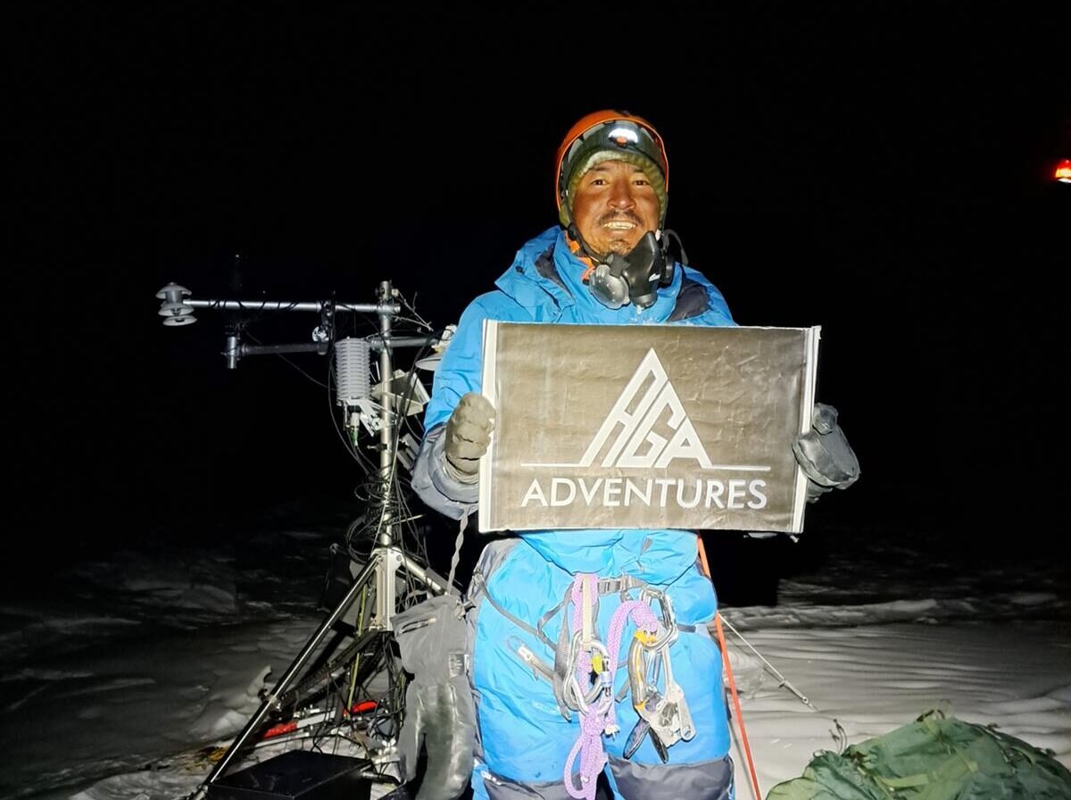 Gelje holds a banner of his company AGA adventures while standing on the flat, snowy summit of Cho Oyu, the meteorological Chinese station behind him, in the middle of the night