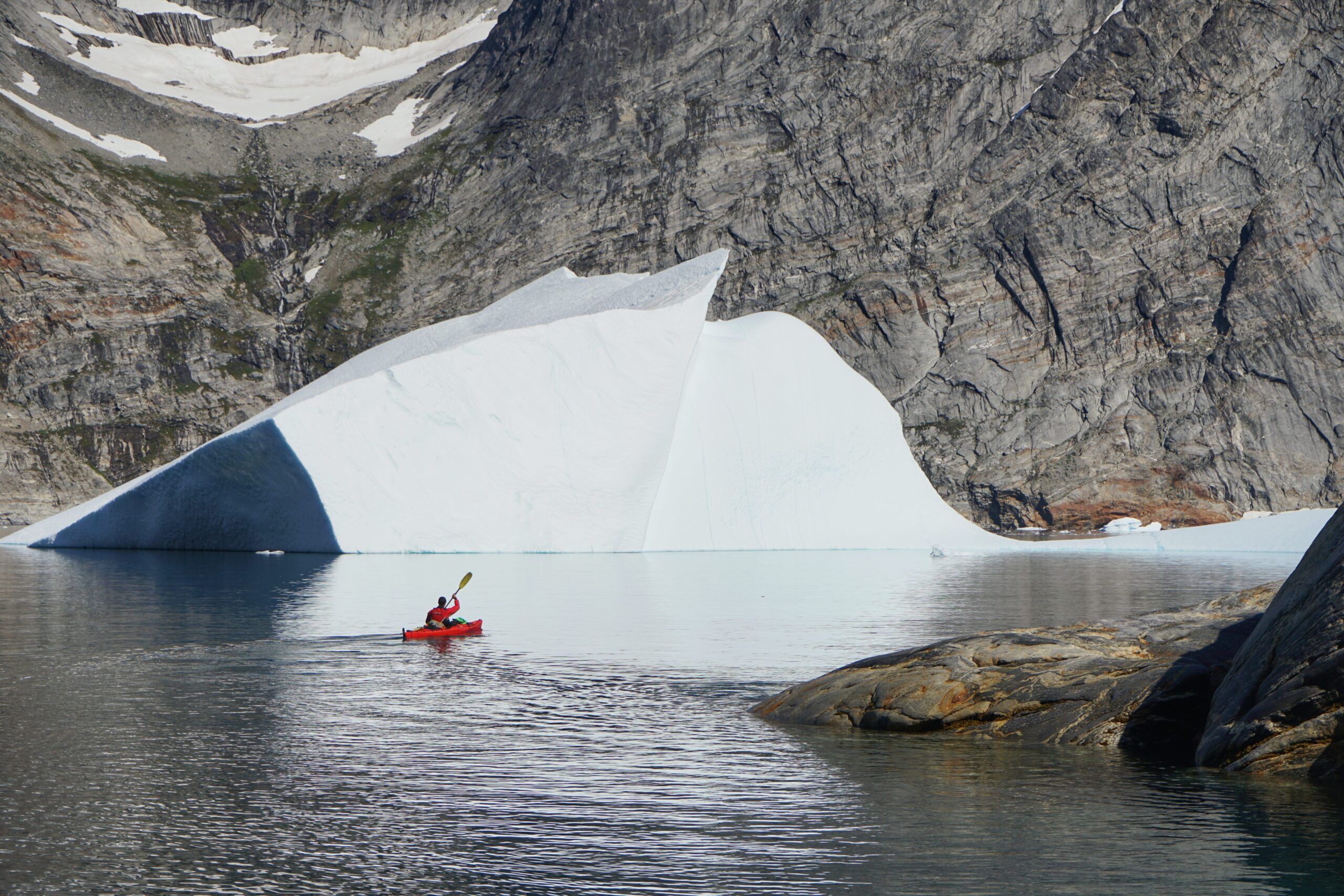 Kayaker in a Greenland fjord with a huge granite wall behind and iceberg