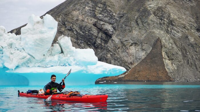 Welfrienger on a kayak, with the rocky fjord and an iceberg behind
