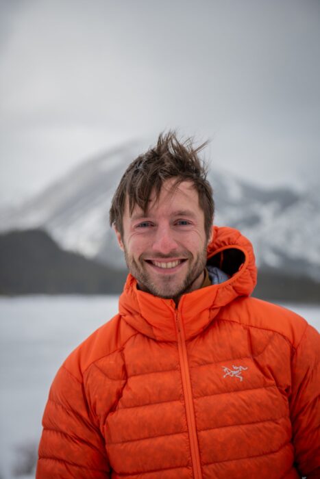 Ethan Berman in orange dawn jacket with a lake and a mountain behind