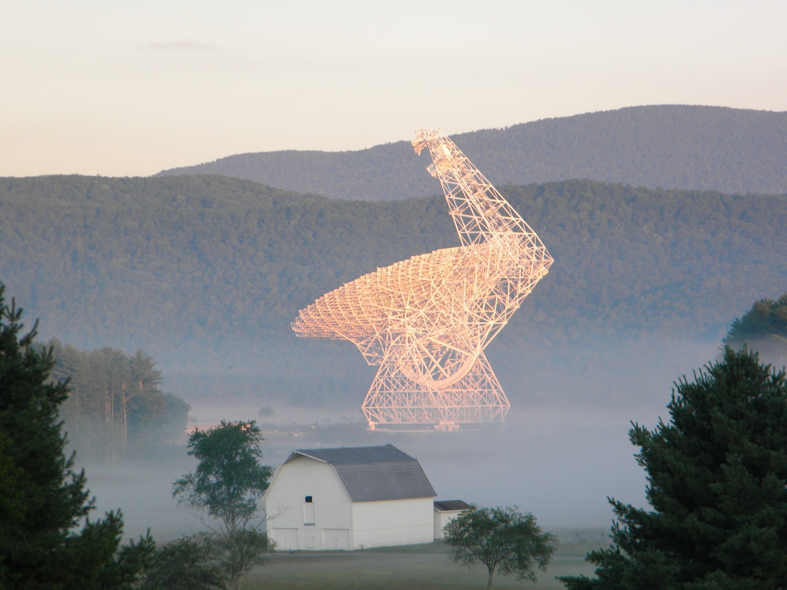 A very large white dish glows in the early morning light, surrounded by mist. A house sits in the foreground; in the background are mountains.