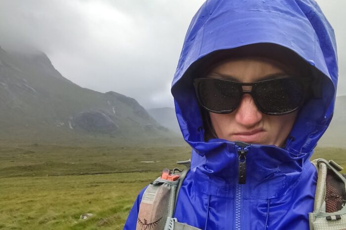 Chelsey is wearing a BD rain jacket that has wetted out, and sunglasses. She's making an annoyed face, and you can see the green hills of Scotland behind her.