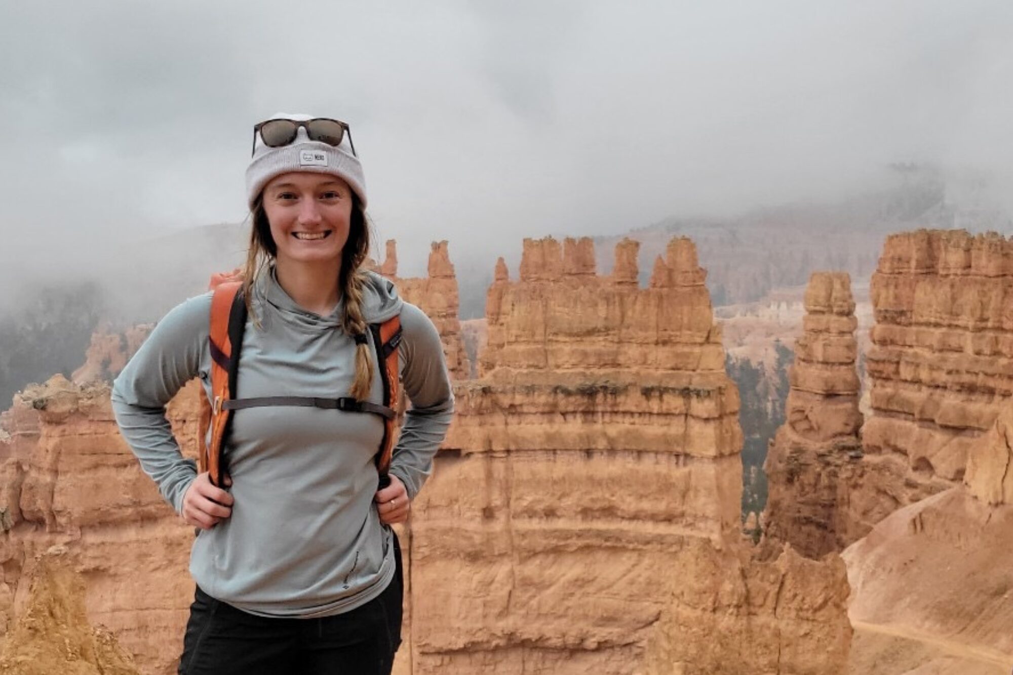 Chelsey is wearing a Black Diamond sun hoody and a grey hat, with an orange backpack. She's standing in front of some misty orange hoodoos in Bryce Natinoal Park.