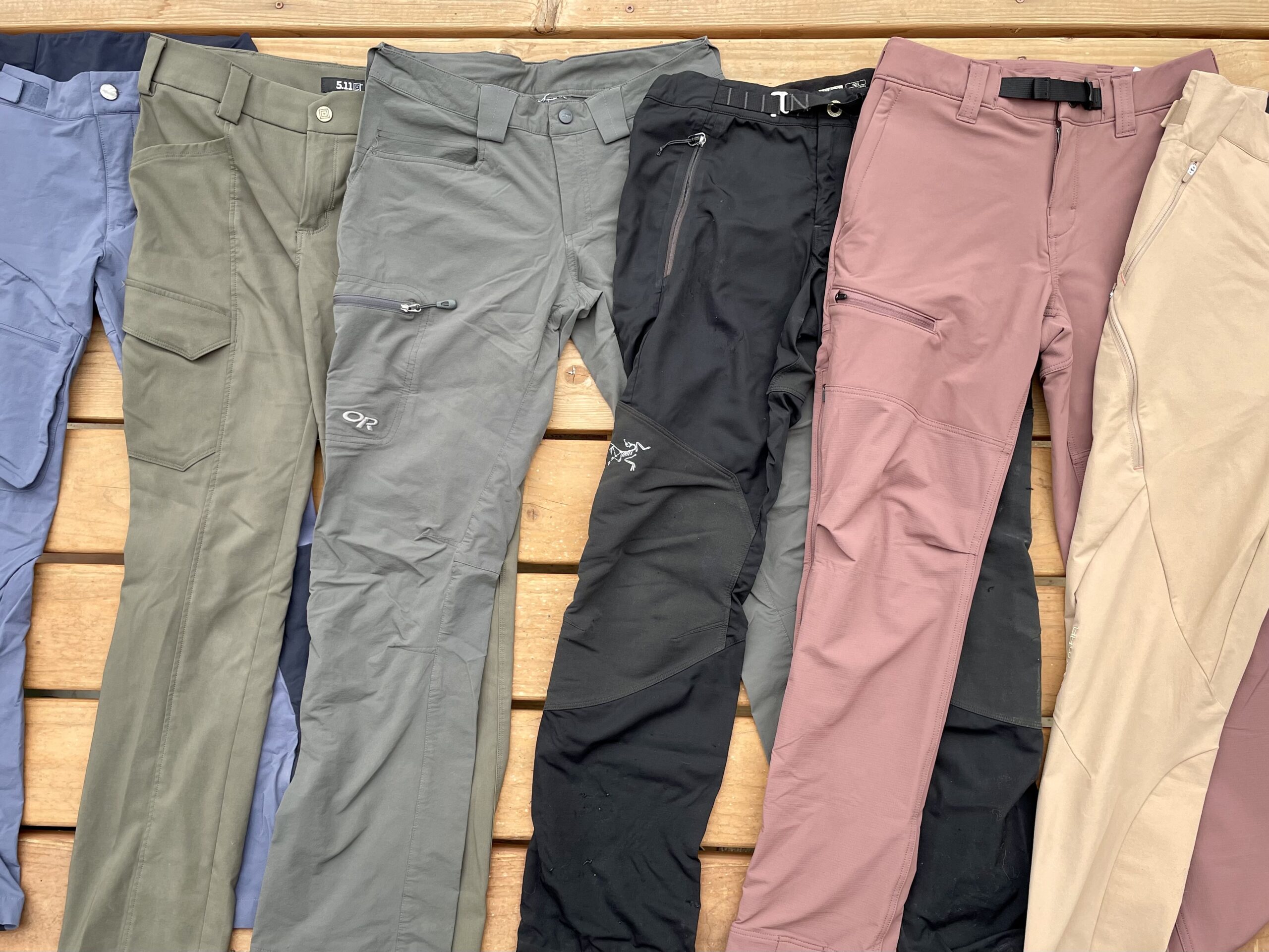 Several pair sof hiking pants are laid on the ground, with blue, green, grey, black, and pink pants overlapping each other.