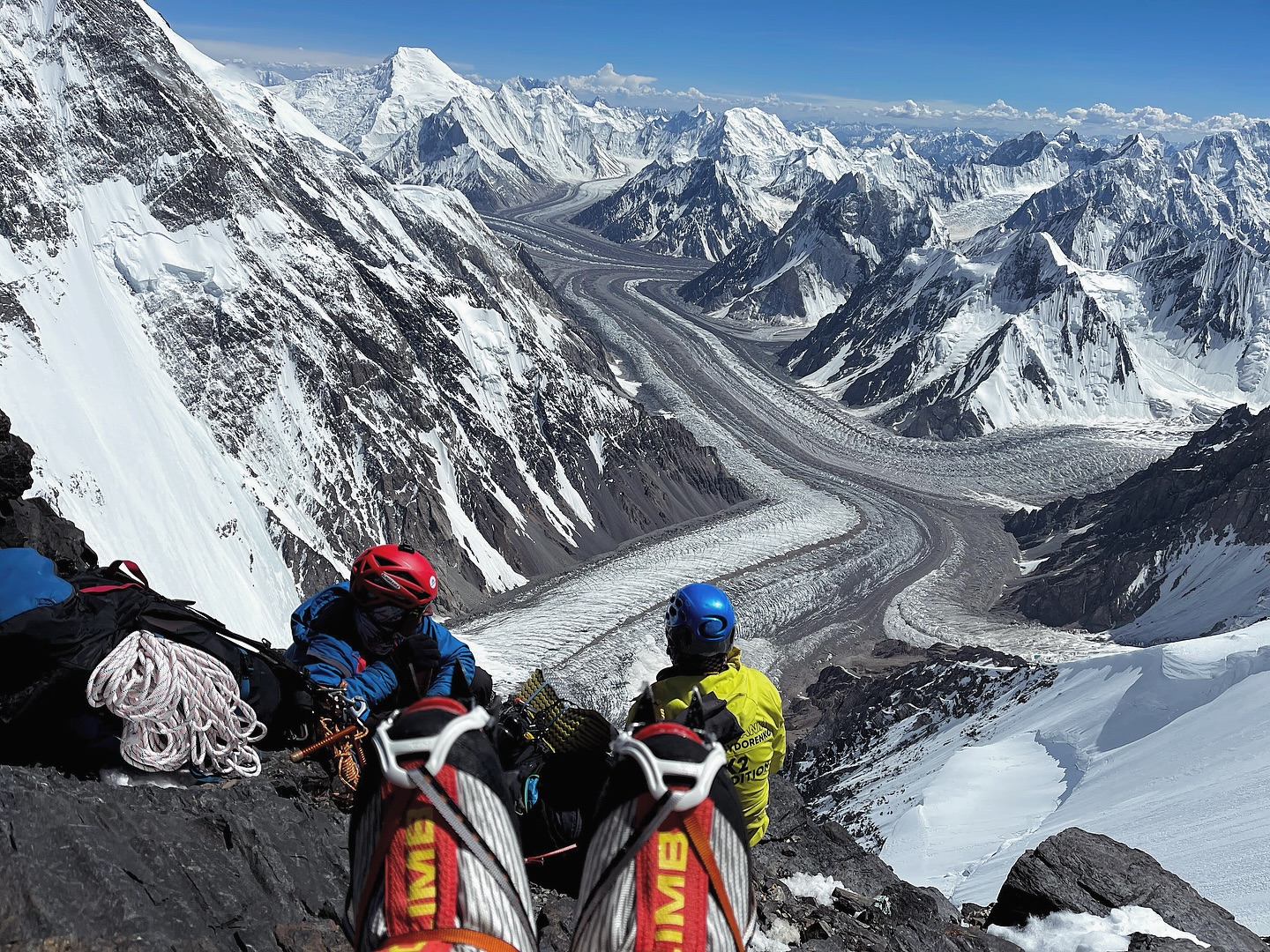 A climber's boots in front, other climbers, the Karakoram glaicers below and Chogolisa in background compose this image shot near Camp 2 on K2.