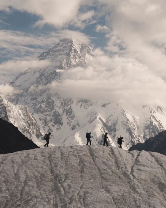 shillouettes of climbers on top of a glacier bump, with K2 behind wrapped in mist