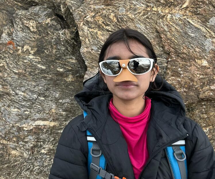 Selena in front of a rock wall, with sunglasses and nose protector.