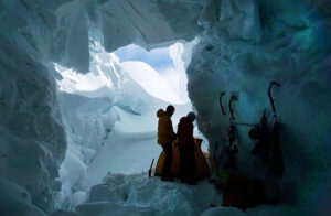 Climbers in a snow cave, seen from the botttom of the cave with seracs and blue sky in background.