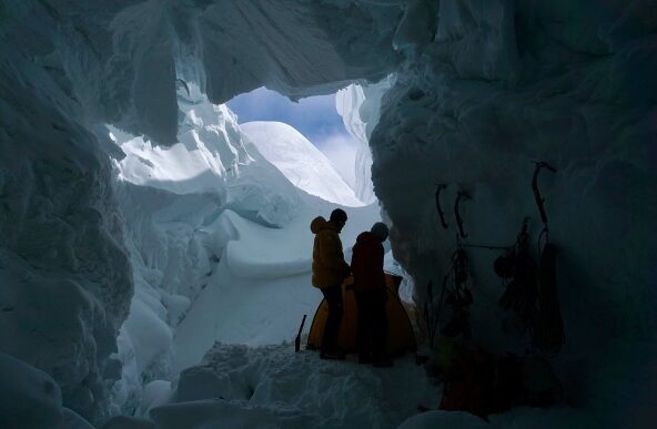 Climbers in a snow cave, seen from the botttom of the cave with seracs and blue sky in background.