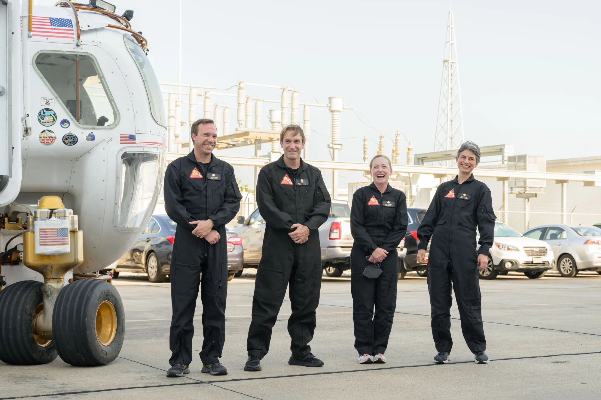 The four crewmembers, dressed in NASA uniforms, stand next to a large rover. Behind them, cars are parked.