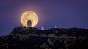 Three telescopes crest a mountain ridge. The full moon silhouettes the largest of them.