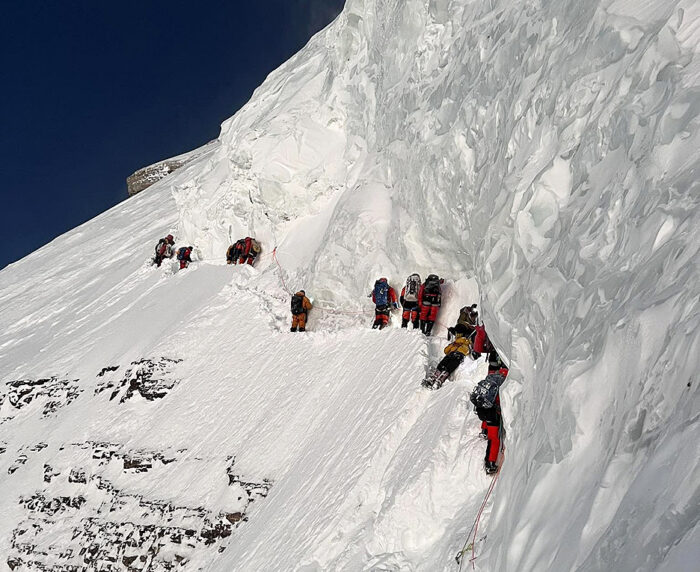 a line of climbers fixed to a rope traverse a snow ramp under a gigantic serac.