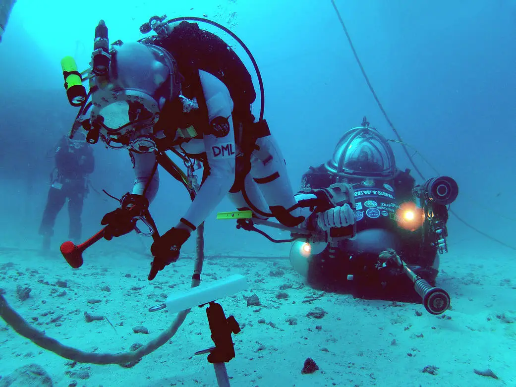 Underwater, an aquanaut in a bulky suit holds a device toward the sea floor. Another aquanaut sits behind them.