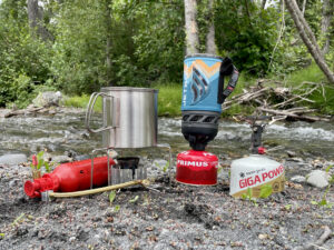 The MSR Dragonfly, Jetboil, and Pocket Rocket Deluxe are set up on a gravel bar by a river.