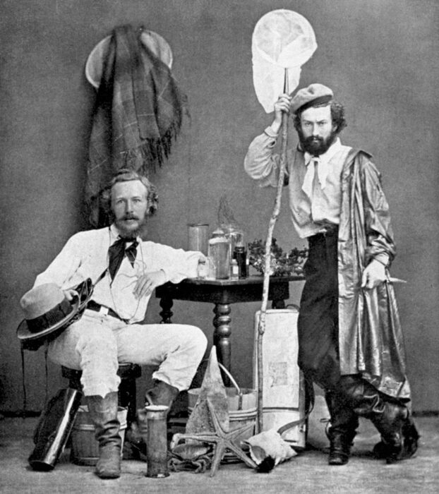 two men in 19th century explorers garb posing for a photo in front of a backdrop