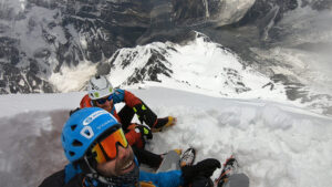 The climbers take a selfie while resting on a snow ledge, the mountain flank at their feet.