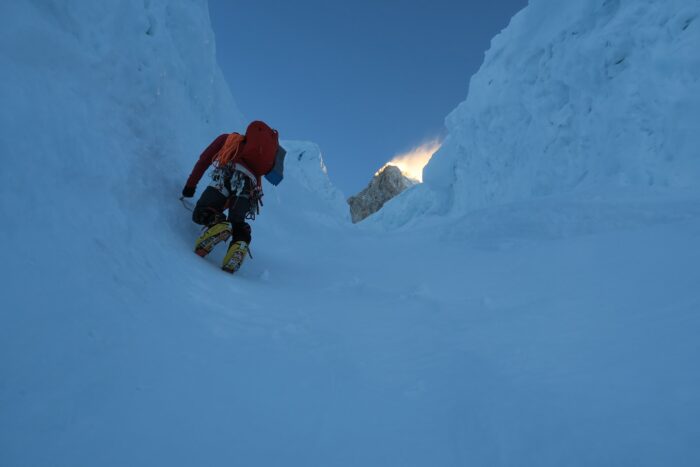 a climber on an ice couloir in the shade, the peak above lit by the sun and hit by he wind