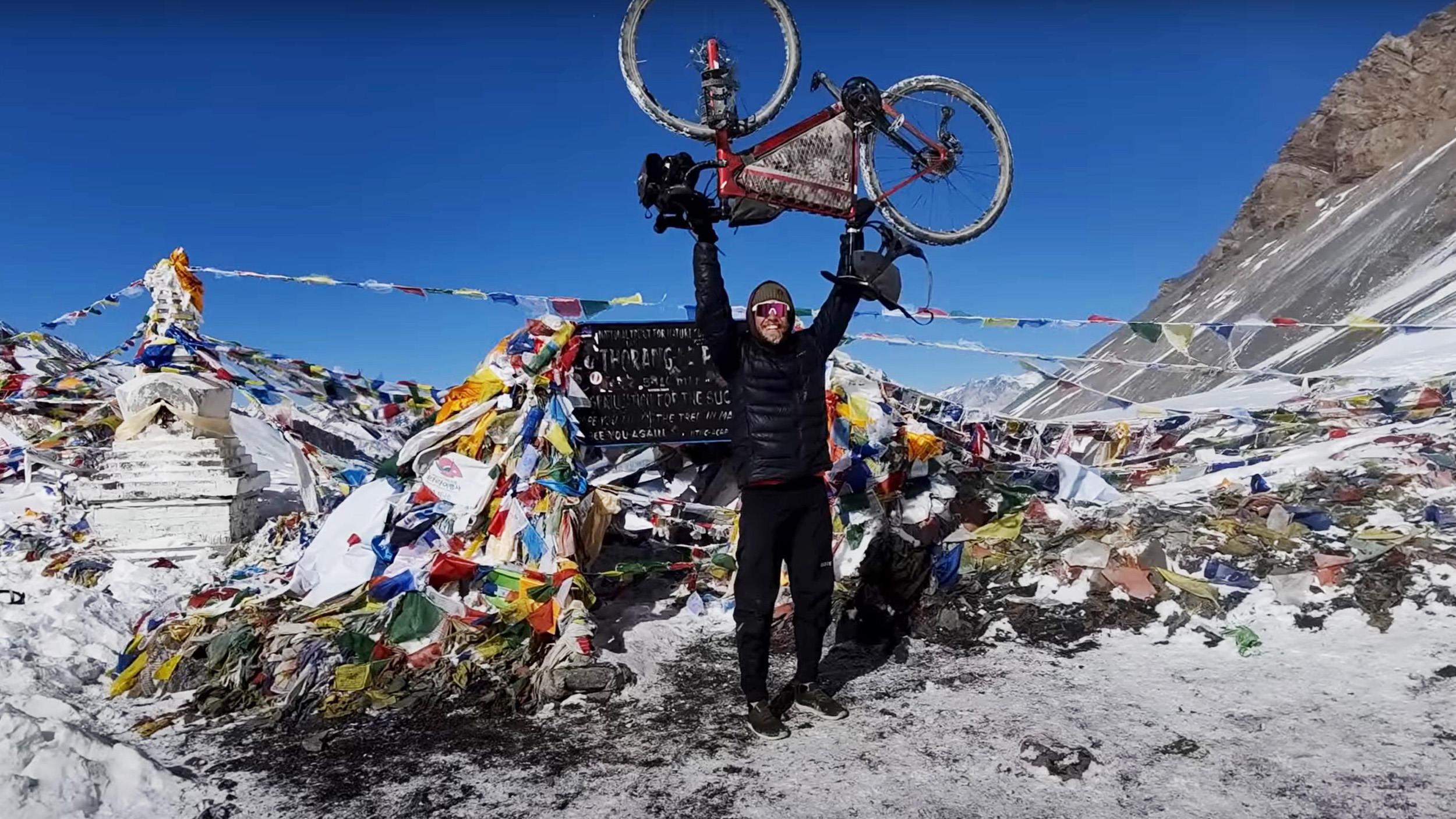 a man holds his bicycle up over his head in front of prayer flags