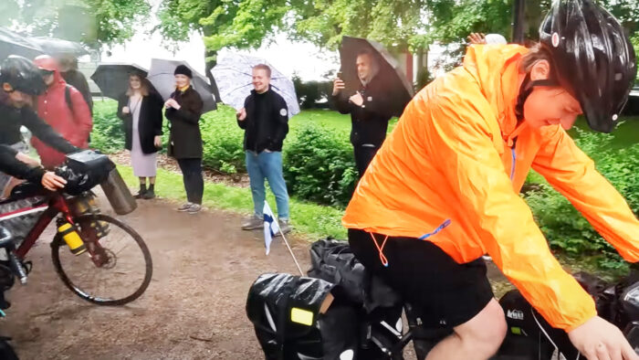 two men ride bicycles surrounded by well wishers seeing them off