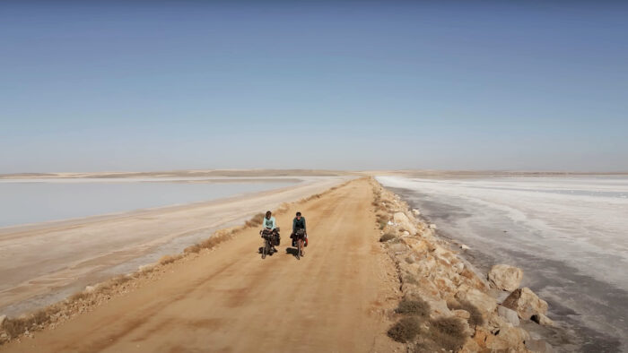 two cyclists ride across a dirt road with salt flats on either side