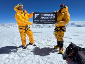 Two Sherpa climbers in yellow dawn suits hold a batter of the flat, snowy summit of Cho Oyu