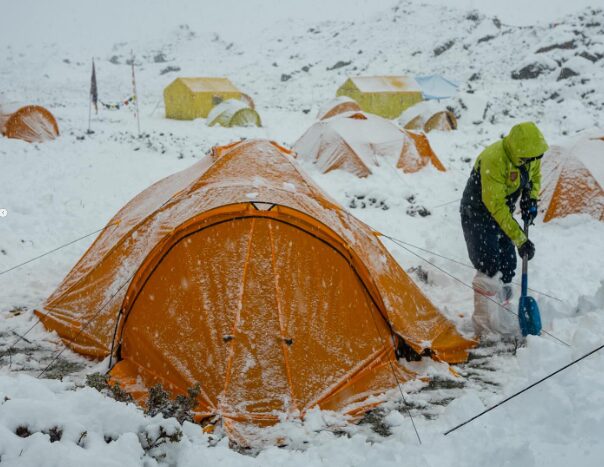 A climber shovels snow away from the gate of a tent in a snow-covered Base Camp