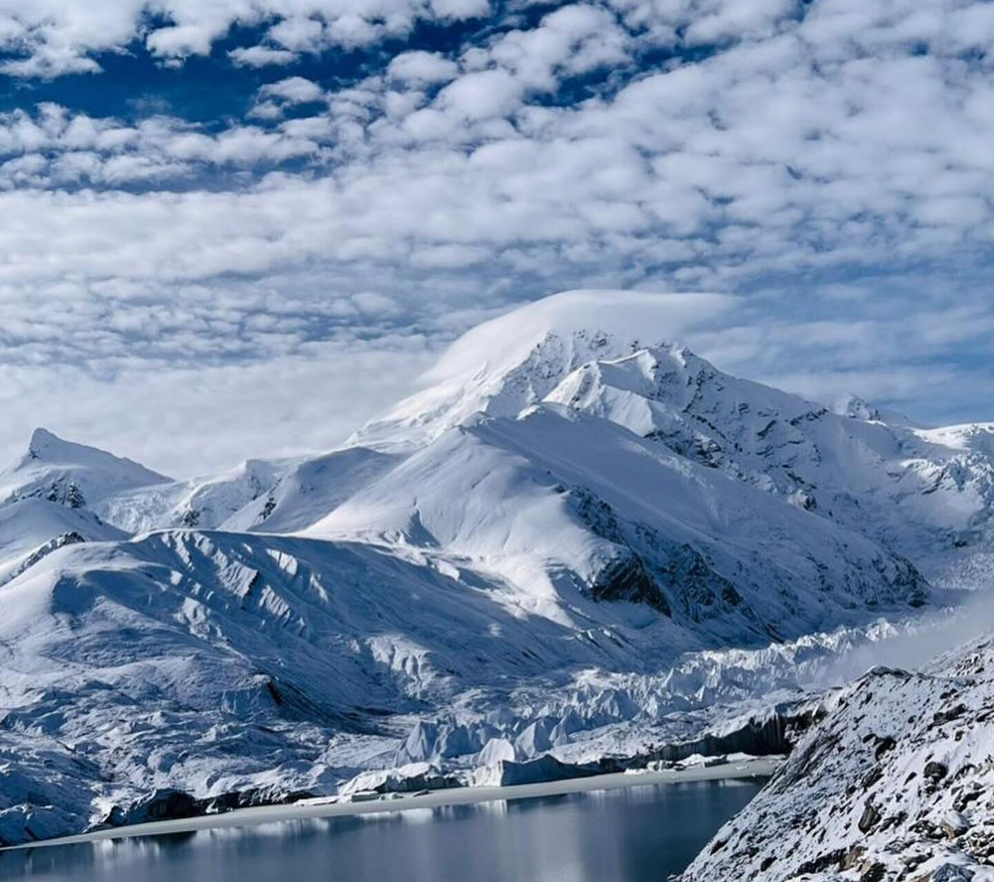 Shisha Pangma under a cloud-dotted sky, a lake below