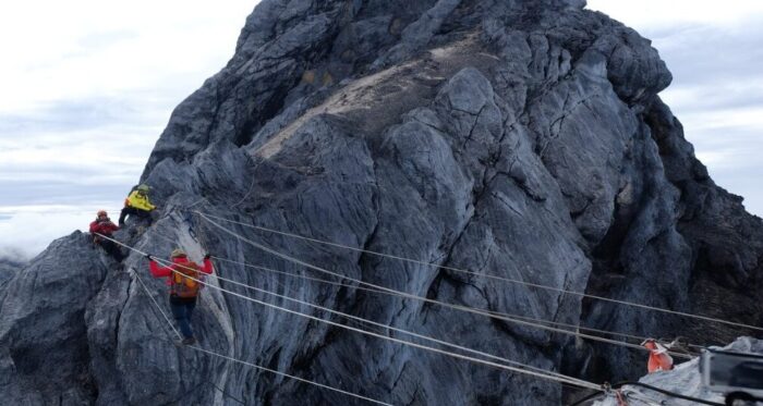 climbers on a cable bridge cross a gap on a rocky peak