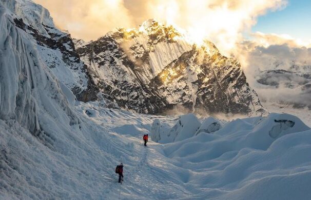 Climbers on a crevased, falt area and a rock and ice face in front of them.