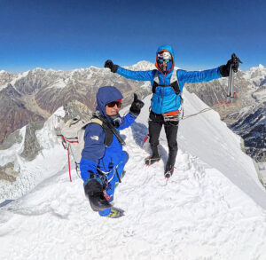 The climbers raise their arms on a narroy, snowy summit in a clear day.