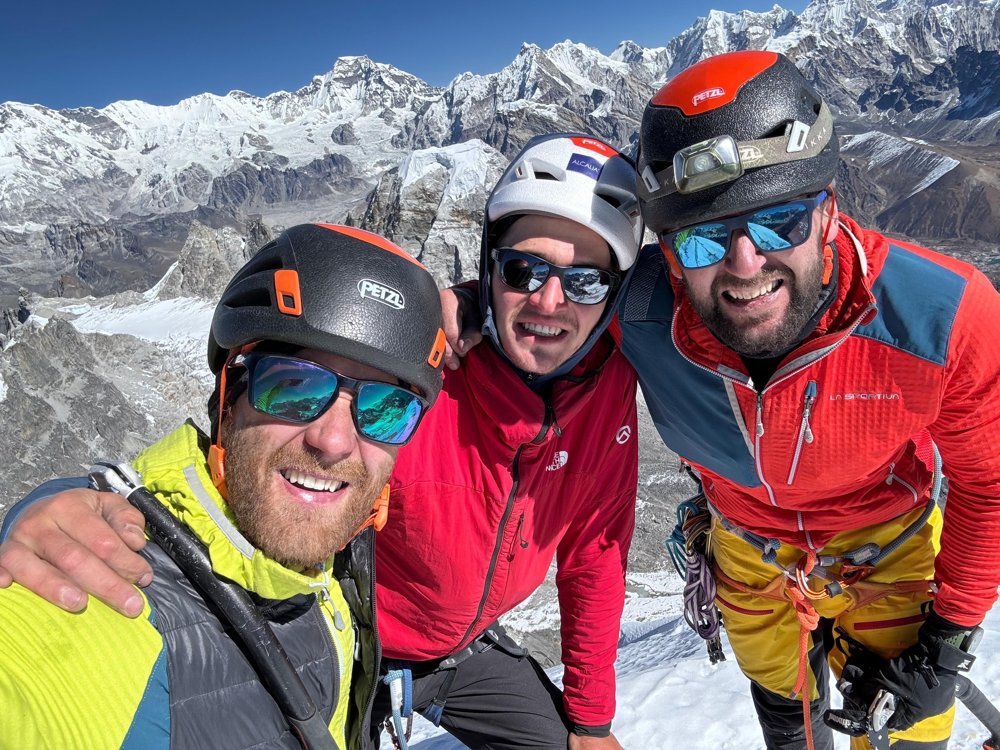 Climbers hold arms and smile on a summit, a horizon of peaks behind them