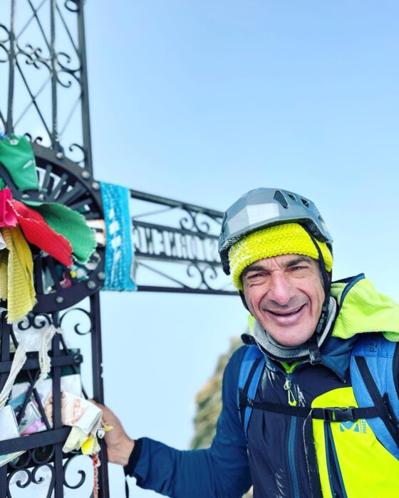 Mattia Conte smiles by a metal cross on a summit. 