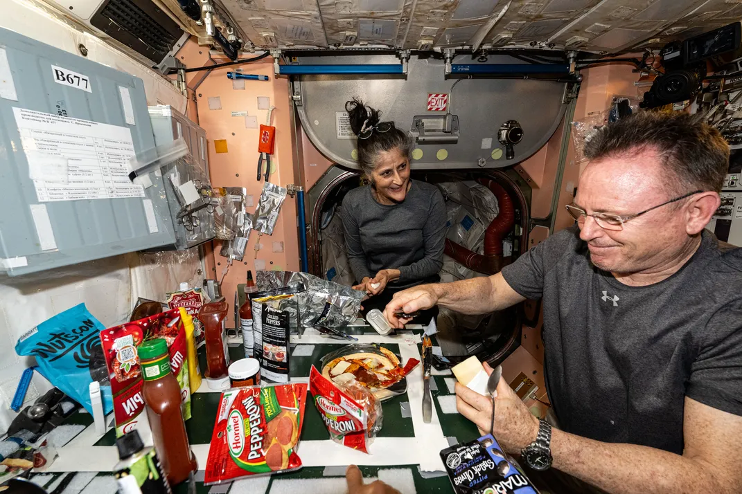 A woman and a man prepare pizza in a cramped room. The angle distorts their faces, making the woman appear stretched and the man squished.